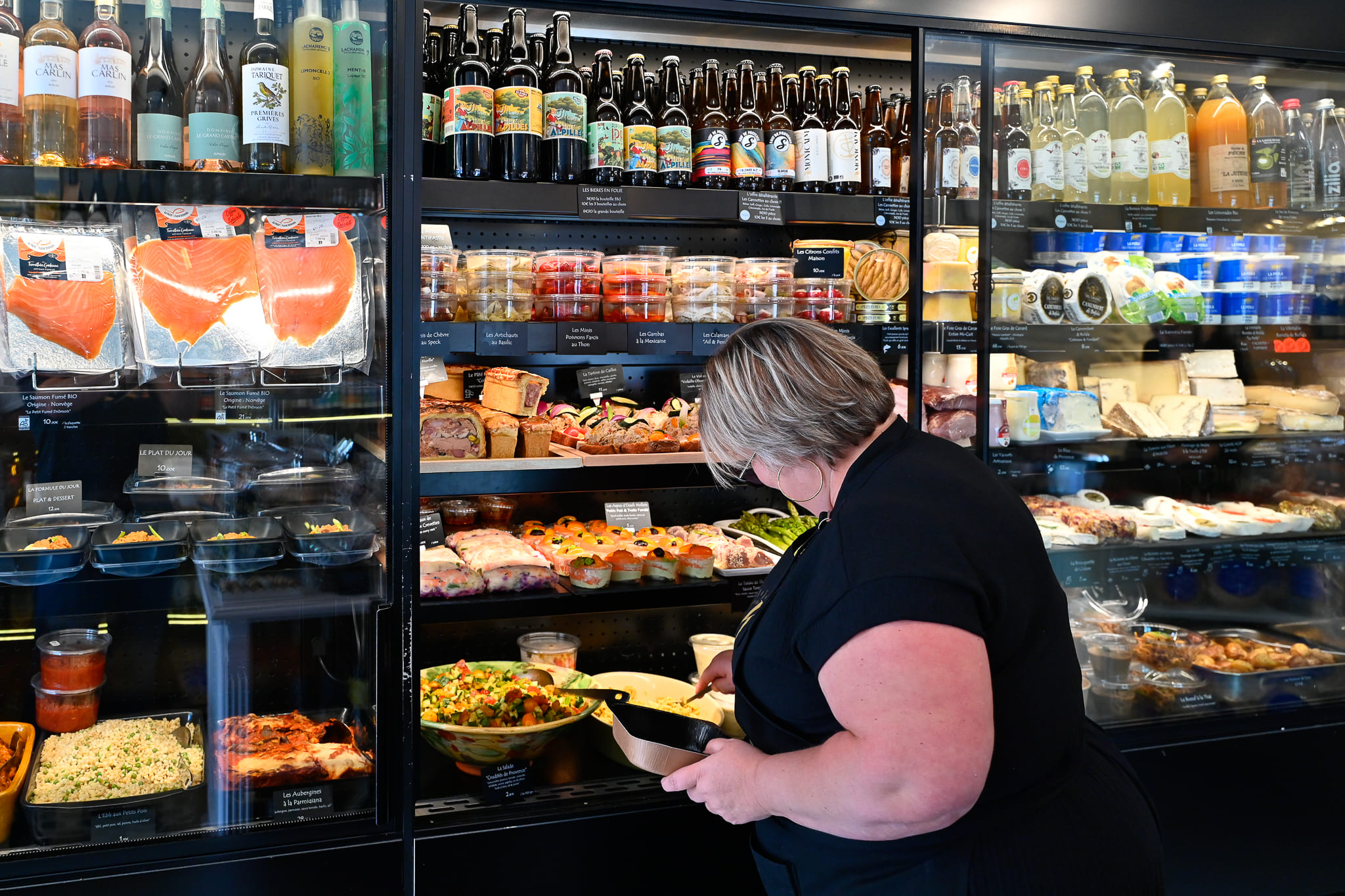 Carte Traiteur Événementiel à Saint-Rémy-de-Provence | Chez Karen Karen vue de profil dans son épicerie fine à Saint-Rémy, sélectionnant des produits devant une grande vitrine réfrigérée traiteur remplie de saumons, antipasti, salades fraîches, fromages et boissons artisanales