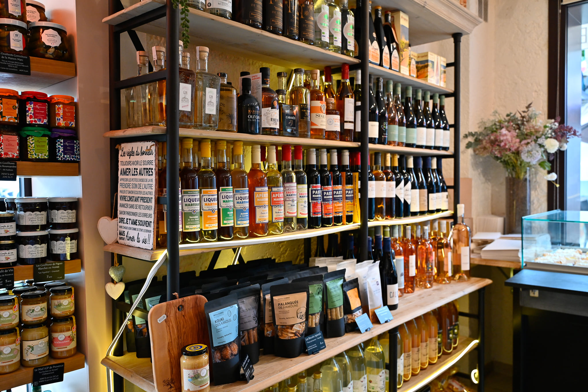 Cave à Spiritueux, Liqueurs et Bières Artisanales – Chez Karen Saint-Rémy Étagères en bois et métal de l'épicerie fine Chez Karen présentant une large sélection de spiritueux locaux (Liqueurs Martinat, pastis, gin), des bouteilles de vin blanc et rosé, ainsi que des biscuits salés apéritifs en sachets au premier plan. N'hésitez pas à prendre contact pour avoir plus d'information.