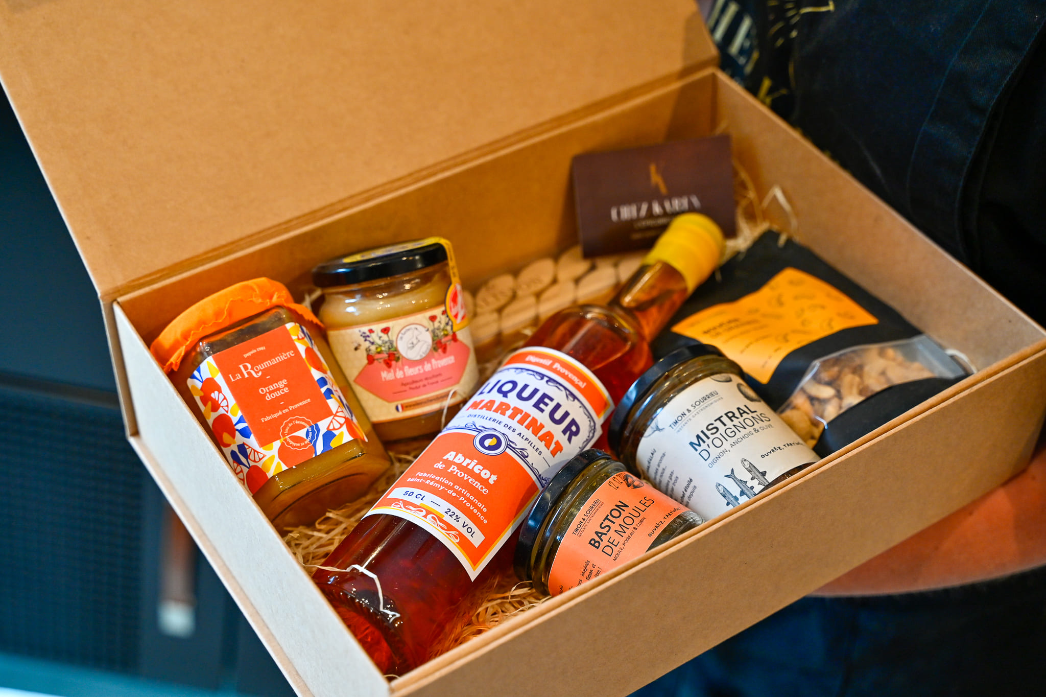 Coffrets Cadeaux Gourmands et Paniers Garnis de Provence – Chez Karen Coffret cadeau en carton kraft ouvert et tenu à la main, garni de pépites locales de Provence : une bouteille de Liqueur Martinat à l'Abricot de Provence, un pot de confiture d'orange douce La Roumanière, du miel de fleurs de Provence, des tartinables Mistral d'Oignons et Baston de Moules, et un sachet de biscuits artisanaux