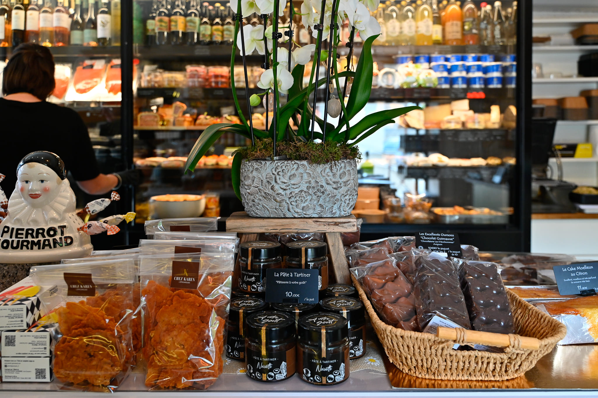 Comptoir d'épicerie fine Chez Karen présentant des sachets de biscuits artisanaux, des pots de pâte à tartiner à la noisette, un panier de chocolats fins et un cake moelleux au citron, le tout disposé sous un pot d'orchidées blanches