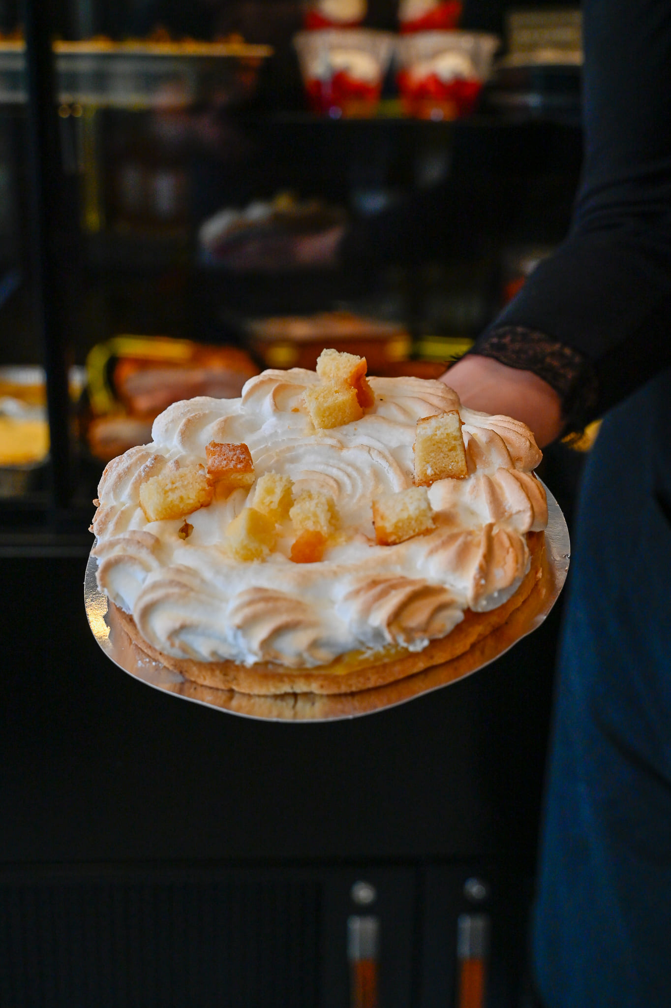Gros plan sur une tarte au citron meringuée artisanale, décorée de petits cubes de génoise moelleuse, présentée sur un support argenté par une personne en boutique.