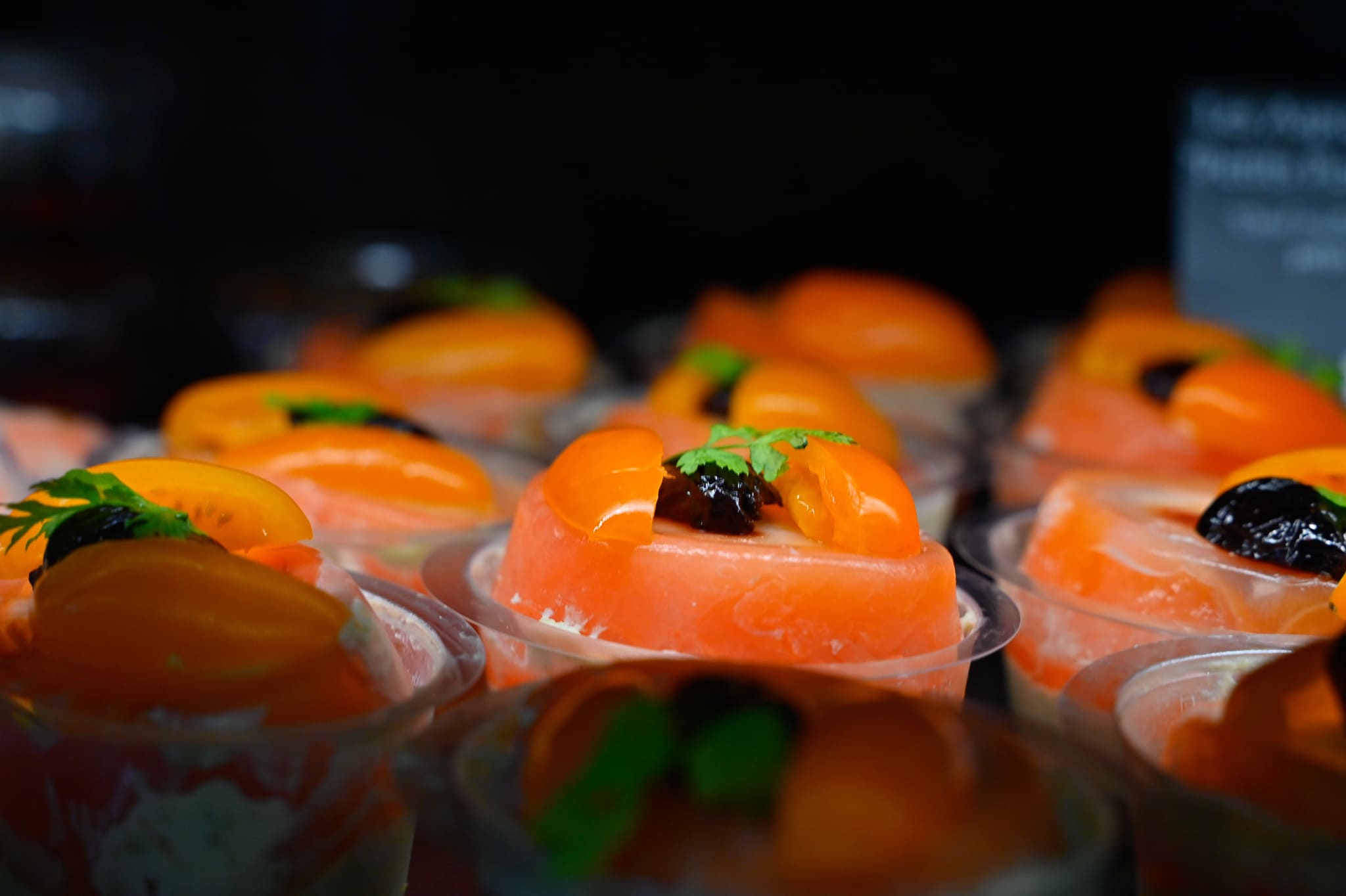 Alignement de verrines traiteur haut de gamme de Chez Karen, décorées de tomates orange éclatantes, d'une touche d'olive noire et d'un brin de cerfeuil frais sur un fond sombre et feutré.