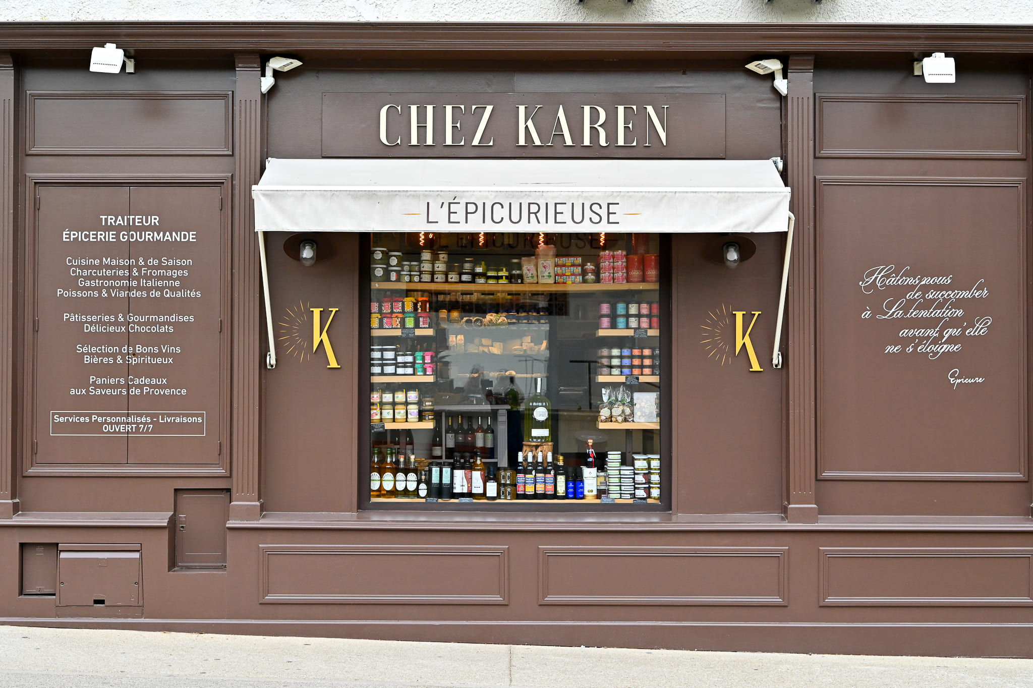 Épicerie Fine et Traiteur L’Épicurieuse à Saint-Rémy-de-Provence Façade en bois marron chocolat de la boutique Chez Karen - L'Épicurieuse à Saint-Rémy-de-Provence, vue de face, révélant ses inscriptions dorées, sa grande vitrine garnie de bocaux et vins, et ses panneaux listant les services traiteur et épicerie gourmande