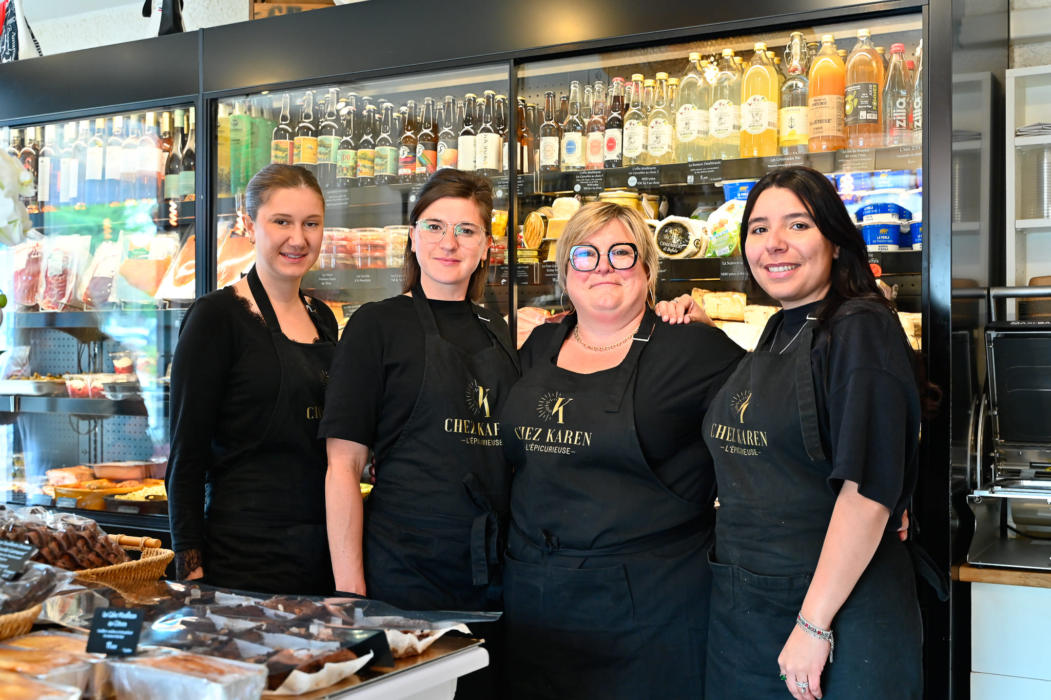 Notre Équipe Passionnée – Épicerie Fine Chez Karen à Saint-Rémy Portrait souriant des quatre membres de l'équipe féminine de Chez Karen L'Épicurieuse, portant des tabliers noirs assortis avec logo doré, réunies dans la boutique devant une grande vitrine traiteur bien garnie.