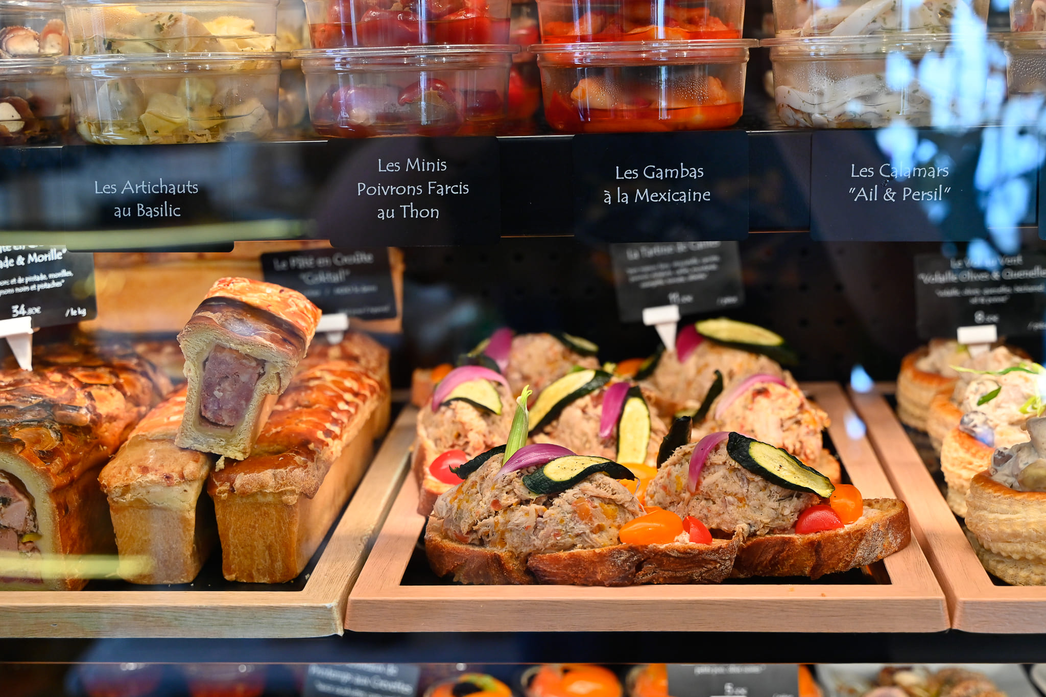 Panier Pique-Nique Chic et Traiteur Terroir – Chez Karen L’Épicurieuse Gros plan sur nos spécialités traiteurs, une vitrine de traiteur Chez Karen présentant du pâté en croûte artisanal, des tartines gourmandes aux crudités (courgettes, oignons rouges) et une sélection d'antipasti en bocaux (artichauts au basilic, poivrons farcis au thon)