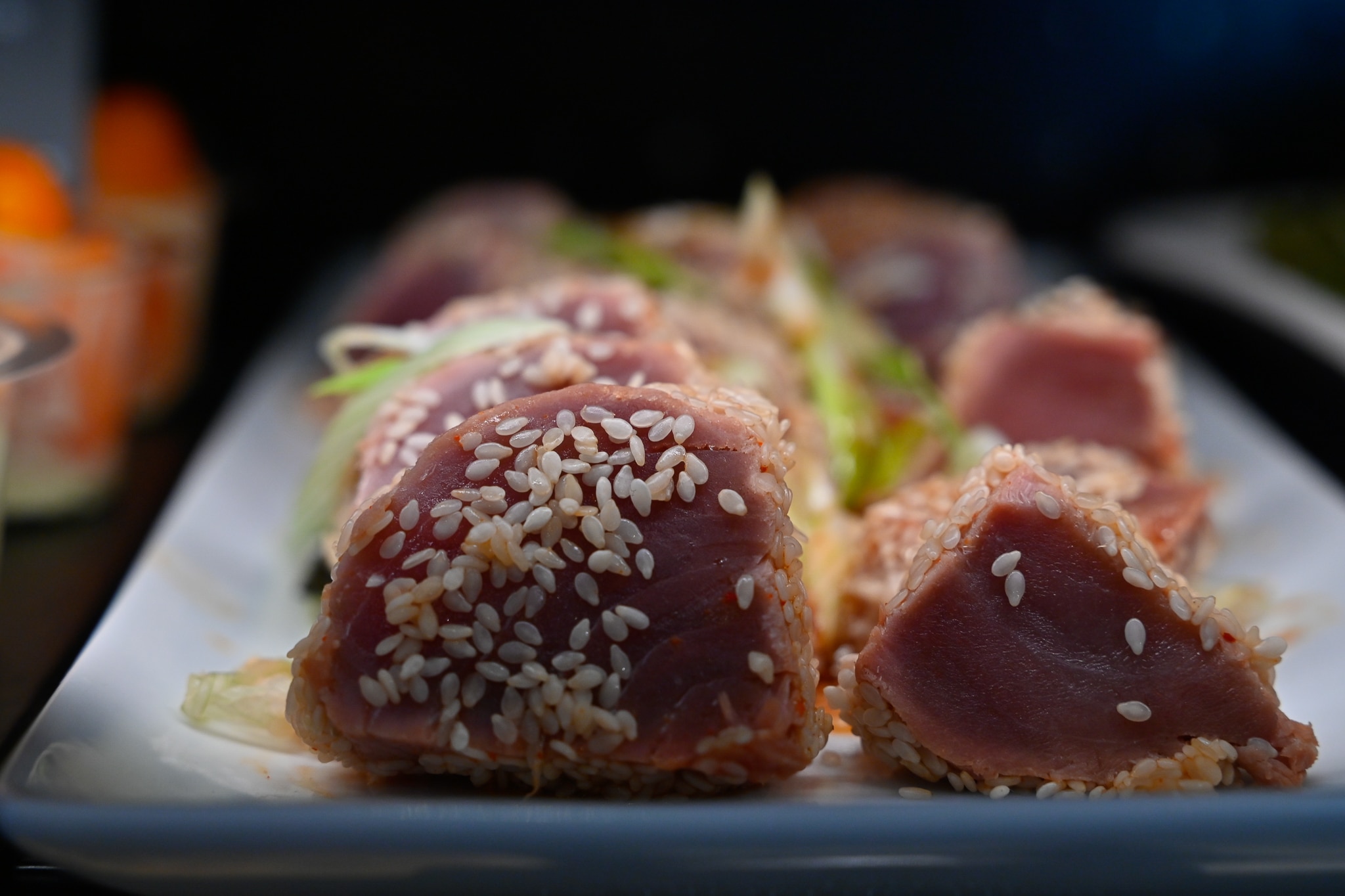 Gros plan sur des tranches de tataki de thon rouge mi-cuit enrobées de graines de sésame blanc, dressées sur un plat blanc avec des oignons nouveaux émincés.