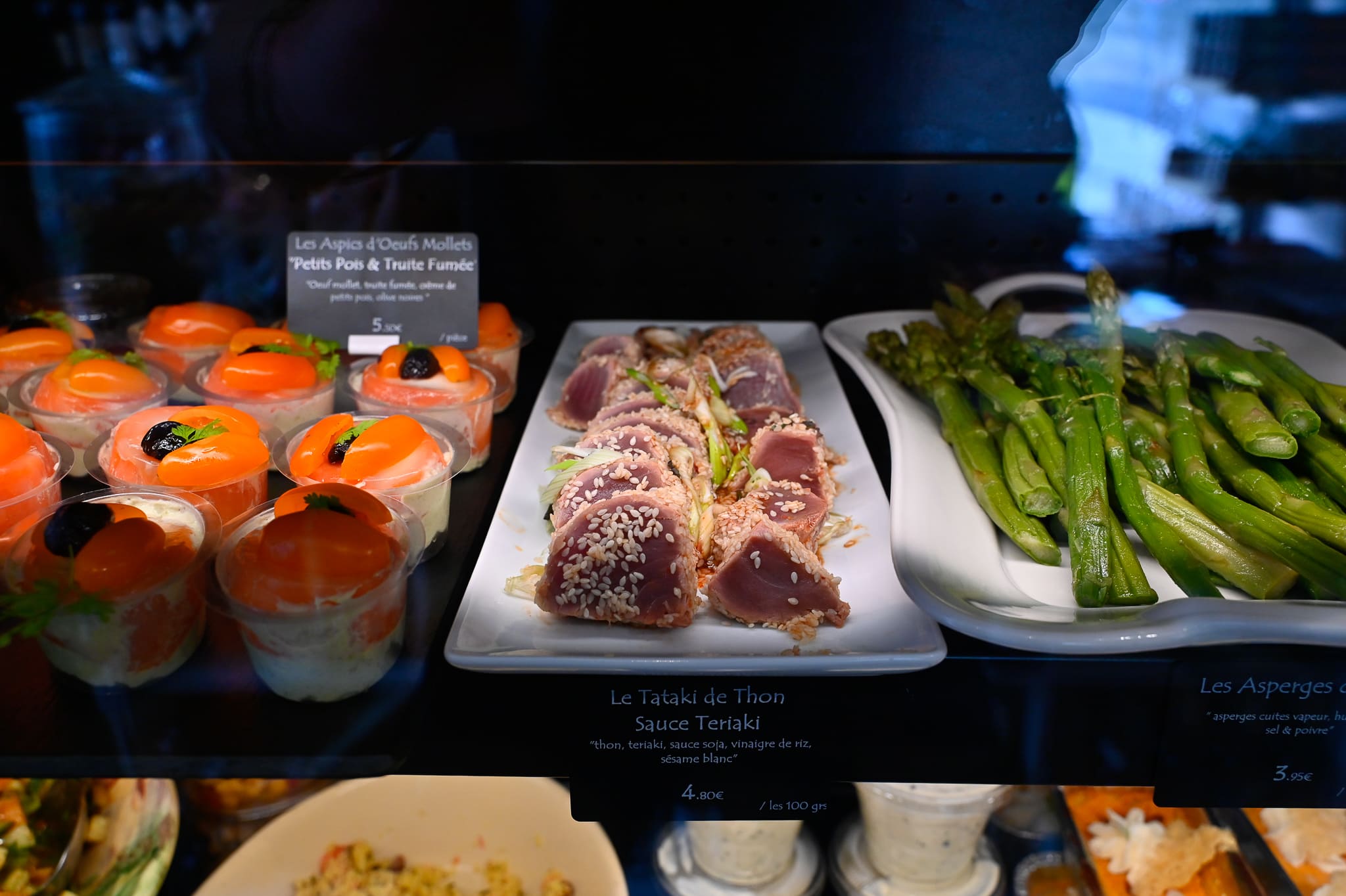 Nos spécialités traiteurs - Vitrine traiteur de l'épicerie fine Chez Karen mettant en valeur un tataki de thon sauce teriyaki au sésame blanc, un plat d'asperges vertes cuites à la vapeur, et des verrines d'aspics d'œufs mollets aux petits pois et truite fumée.