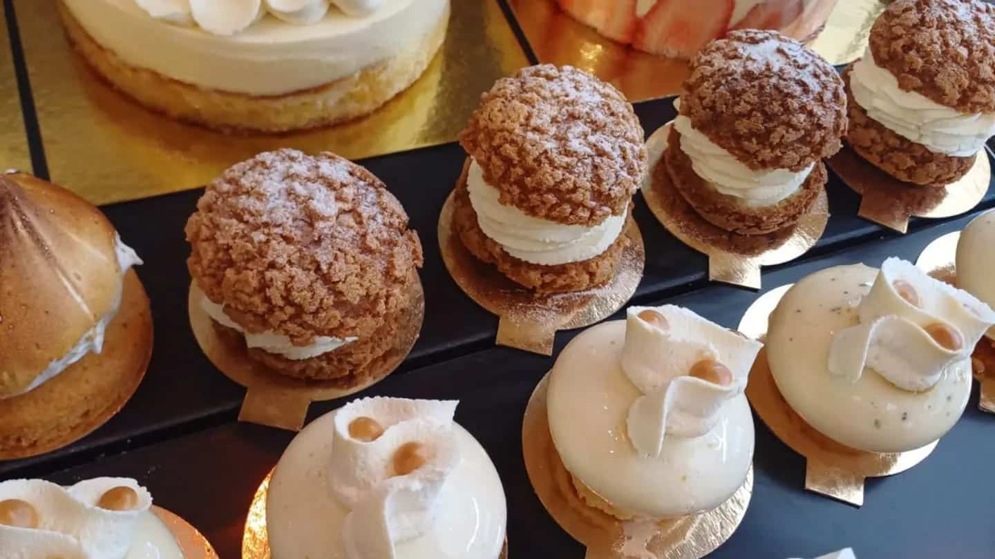 Alignement de pâtisseries individuelles artisanales : des choux à la crème avec craquelin croustillant et sucre glace, et des entremets blancs brillants avec décors élégants sur des ronds dorés.